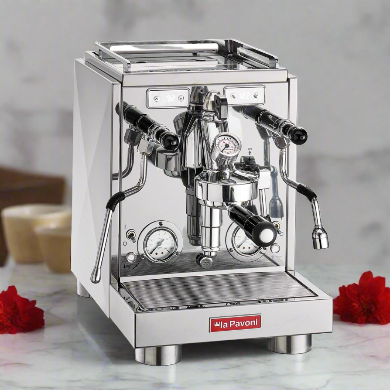 La Pavoni Botticelli Specialty Dual Boiler Coffee Machine in polished stainless steel, featuring dual pressure gauges, steam wand, hot water spout, and premium portafilter. Contemporary design with red brand badge, displayed on marble counter with red flower accents.