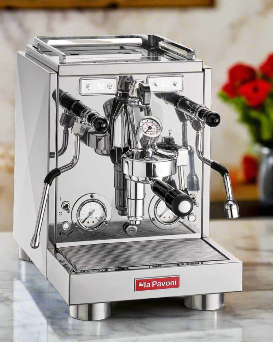 La Pavoni Botticelli Specialty Dual Boiler Coffee Machine in polished stainless steel, featuring professional-grade dual pressure gauges, steam wand, hot water spout, and drip tray. Premium Italian espresso machine photographed on marble countertop with red flowers in background.